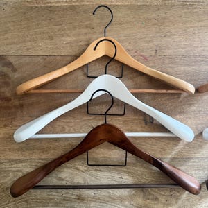 May include: Three wooden clothes hangers with black metal hooks and horizontal bars. The top hanger is light natural wood, the middle hanger is painted white, and the bottom hanger is a dark stained wood finish, all featuring curved shoulder shapes.