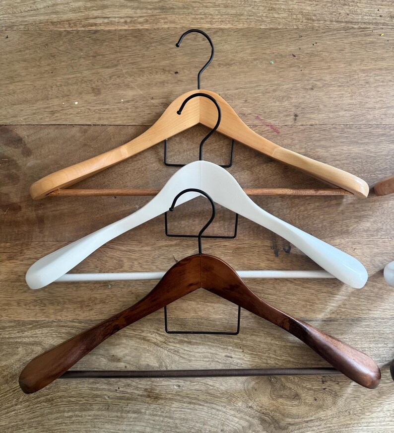 May include: Three wooden clothes hangers with black metal hooks and horizontal bars. The top hanger is light natural wood, the middle hanger is painted white, and the bottom hanger is a dark stained wood finish, all featuring curved shoulder shapes.