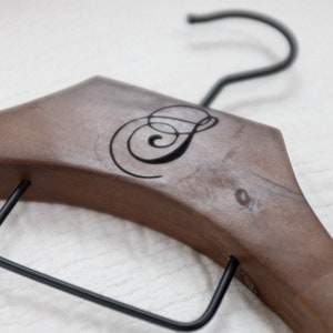 May include: A brown wooden clothes hanger featuring a black metal hook and a black metal bar across the bottom. The center of the wooden frame is engraved with a stylized black letter S in a cursive script font, centered on the smooth wood surface.