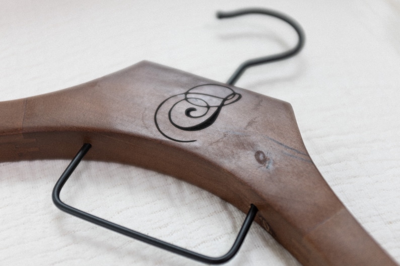May include: A brown wooden clothes hanger featuring a black metal hook and a black metal bar across the bottom. The center of the wooden frame is engraved with a stylized black letter S in a cursive script font, centered on the smooth wood surface.