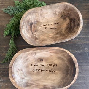 Puede incluir: Dos cuencos de madera de forma ovalada con un acabado rústico. Los cuencos tienen inscripciones manuscritas: "I love you Daddy Evy-Grace" y "I love you Daddy Weston". La vegetación acentúa la escena, añadiendo un toque natural.