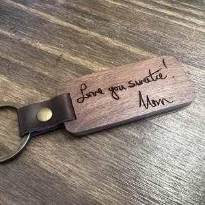 May include: A rectangular wooden keychain with a leather strap and metal ring. The keychain is engraved with the words "Love you sweetie! Mom" in a handwritten style. The wood has a natural grain.