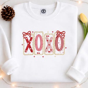 May include: White sweatshirt with embroidered "XOXO" design. The letters are in red and pink, with heart and bow accents. The design is framed with a gold border. A pinecone and a yellow tulip are visible in the background.