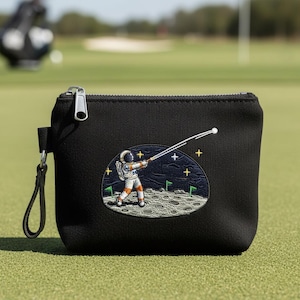 May include: Black zippered pouch with a whimsical embroidered design of an astronaut golfing on the moon. The astronaut is wearing a spacesuit and swinging a golf club. The pouch has a small loop for easy carrying.