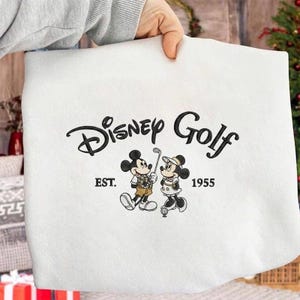 Könnte beinhalten: Weißes Sweatshirt mit dem Logo "Disney Golf" in schwarzer Schrift, mit Mickey und Minnie Mouse in Golfkleidung. Der Text "EST. 1955" befindet sich unter dem Logo. Das Sweatshirt wird von einer Person gehalten.