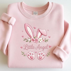 Può includere: Felpa rosa chiaro con un disegno ricamato di orecchie di coniglio, fiori, cuori e impronte di zampe. Il testo "Little Angel" è anch'esso ricamato. Girocollo e maniche con risvolto.
