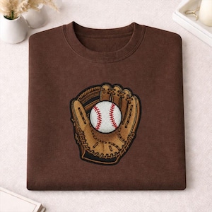 May include: A brown sweatshirt with an embroidered baseball and mitt design. The baseball is white with red stitching, and the mitt is tan and brown. The sweatshirt has a crew neck.