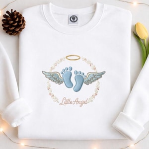 May include: White sweatshirt with an embroidered design featuring baby footprints, wings, and a halo. The words "Little Angel" are embroidered in a delicate script. A pinecone and a yellow tulip are visible in the background.
