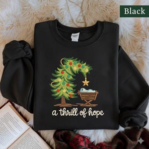 Puede incluir: Sudadera negra con un diseño de árbol de Navidad. El árbol verde está decorado con adornos rojos y amarillos, una estrella dorada y un bebé en un pesebre. El texto "a thrill of hope" está debajo.