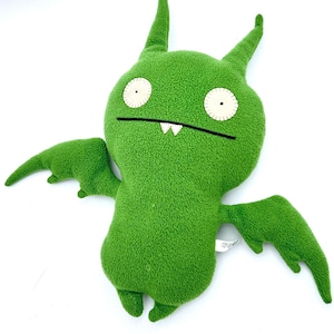 May include: A bright green plush toy shaped like a bat with two pointed ears, two large circular white eyes with black pupils, and a wide black mouth featuring two small white teeth. The creature has two outstretched wings and two small feet.