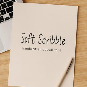 May include: A cream-coloured sheet of paper with the words "Soft Scribble" and "handwritten casual font" in black cursive. A light-coloured pen rests on the paper, and a laptop is partially visible in the background.