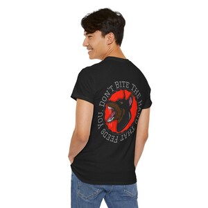 以下が含まれることがあります： 赤い円の中にドーベルマンピンシャーのグラフィックが描かれた黒いTシャツ。「YOU DON'T BITE THE HAND THAT FEEDS YOU」の文字が入っています。犬は口を開けて歯が見えるように描かれています。