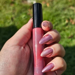 May include: A pink lip gloss tube with a black cap is held in a hand. The lip gloss has a gradient effect, transitioning from a darker pink at the base to a lighter shade towards the top. The person's nails are painted a metallic purple.