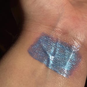 May include: A swatch of iridescent blue makeup on a person's skin. The makeup has a shimmering, metallic finish with hints of silver and purple. The texture appears smooth and creamy, reflecting light.