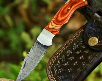Puede incluir: Un cuchillo plegable con una hoja de acero estampada y un mango de madera. El mango es de color marrón rojizo con remaches de latón. El cuchillo está junto a una funda de cuero marrón con detalles en relieve.