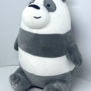Op de afbeelding: Een pluche pandabeer met een witte kop, donkergrijze oogvlekken, een zwarte neus en een geborduurde glimlach. Het lichaam heeft horizontale strepen van witte en donkergrijze pluche stof, met donkergrijze voeten en zooltjes.
