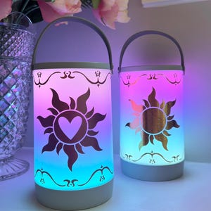 May include: Two cylindrical lanterns with a handle, featuring a sun design with a heart in the center. The lanterns have a light gray base and handle, with a decorative border. The lanterns glow with a gradient of colors, including pink, blue, and purple.