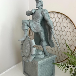 May include: A grey statue of a man in a cape, standing on a pedestal. The statue has a textured surface and is holding a sword. The pedestal has a decorative emblem. The statue is approximately 30 cm tall.
