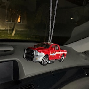 May include: A red and silver toy pickup truck with black wheels and a black string for hanging. The truck has the words "UTILITY 1" on the side. The truck is hanging from a car's rearview mirror.