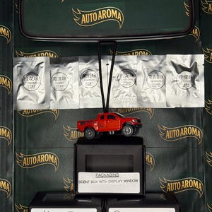 May include: A red toy pickup truck car air freshener suspended from a rearview mirror. The packaging includes scent sachets and boxes with the "AUTO AROMA" logo. The text "SCENT BOX WITH DISPLAY WINDOW" is visible.