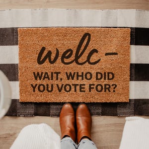 May include: A rectangular doormat with the word "welc-" and the phrase "WAIT, WHO DID YOU VOTE FOR?" in black lettering. The mat is brown and sits on a black and white checkered rug. Brown boots and blue jeans are visible.