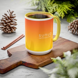 May include: A ceramic coffee mug with a gradient design, transitioning from orange to yellow, with a white base and yellow handle. The mug contains dark liquid and features the text "SUNUP IMPRESSIONS". The mug is on a wooden cutting board.