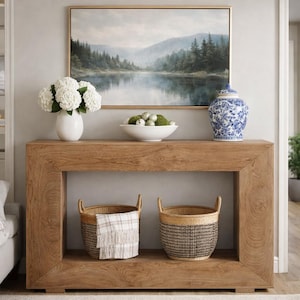 May include: A wooden console table with a framed landscape painting above it. The table displays a white vase with white flowers, a white bowl with decorative spheres, and a blue and white patterned jar. Two woven baskets sit below.