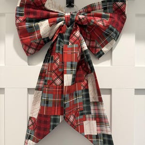 May include: A large decorative bow made from fabric featuring a patchwork pattern of red, green, and white plaid squares. The bow has two wide loops at the top and two long, pointed tails hanging down, all constructed from the same tartan-inspired textile.