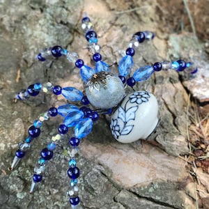May include: A decorative spider ornament featuring a round, speckled grey bead for the head and a white ceramic bead with a blue floral pattern for the abdomen. Eight legs extend outward, crafted from a mix of dark blue, light blue, and clear beads.