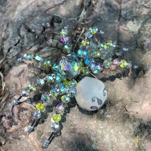 May include: A decorative spider figurine crafted with iridescent, multi-colored faceted crystal beads forming the legs and head, paired with a smooth, round, off-white stone-like abdomen featuring subtle, dark, swirling decorative markings.