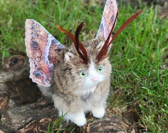 Puede incluir: Una figura de gato artesanal con alas de mariposa y detalles de plumas. El gato tiene pelaje marrón y blanco, ojos verdes y nariz rosa. Las alas presentan un diseño detallado en tonos marrones, naranjas y blancos. La figura se encuentra sobre hierba verde.