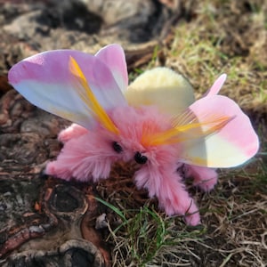 May include: A plush, pink moth toy with feathery wings in shades of pink, white, and yellow. The moth has small black eyes and yellow antennae. The toy is set against a backdrop of grass and wood.