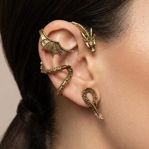 Può includere: Set di gioielli per orecchie a tema drago color oro. Il set include una testa di drago, un ear cuff ad ala, un avvolgimento per l'orecchio a forma di serpente e un orecchino a bottone a forma di piuma. I gioielli sono indossati sull'orecchio.