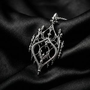 May include: A silver pendant necklace with an ornate, gothic-inspired design. The pendant features a detailed, openwork pattern with pointed arches and delicate filigree. The necklace is displayed on a black satin background.