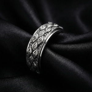 Może przedstawiać: Srebrny pierścionek z misternym wzorem w kształcie oczu. Pierścionek wykonany jest z błyszczącego metalu i ma szczegółową, teksturowaną powierzchnię. Pierścionek jest prezentowany na czarnej, jedwabistej tkaninie, podkreślającej jego właściwości odbijające światło.