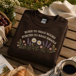 May include: A brown sweatshirt with the embroidered text "BORN TO READ BOOKS FORCED TO READ EMAILS" above a floral design featuring daisies, lavender, and other flowers. The sweatshirt is displayed on a wooden table with a cup of coffee and a basket of grapes.