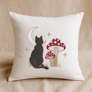 Könnte beinhalten: Cremefarbenes Zierkissen mit einem gestickten Design einer schwarzen Katze, einem Mond, Sternen und zwei rot-weiß gepunkteten Pilzen. Das Kissen hat ein weiches, strukturiertes Aussehen.