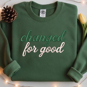 May include: A dark green sweatshirt with the words "changed for good" embroidered in a two-tone green and white script. A pinecone and a yellow tulip are visible in the background.
