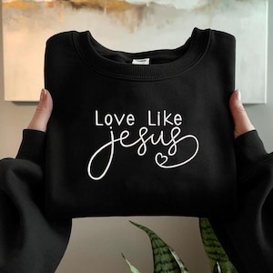 May include: Black sweatshirt with the text "Love Like Jesus" in white script. The word "Jesus" is written in a flowing cursive font, with a small heart symbol. The sweatshirt is a solid black color.