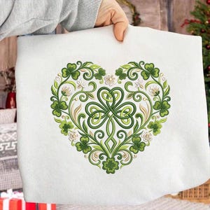 Può includere: Articolo in tessuto bianco con un design a forma di cuore. Il design presenta trifogli verdi, intricati motivi floreali verdi e oro e piccoli fiori bianchi. Il design è centrato.