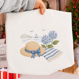 Puede incluir: Un bolso tote blanco con un bordado de escena de playa. El diseño incluye un sombrero de paja con una cinta azul, hortensias azules, una toalla a rayas y un velero en el agua. El bolso es sostenido por una persona.