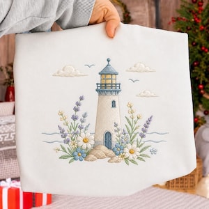 Puede incluir: Funda de almohada blanca con un bordado de faro. El diseño presenta un faro azul claro y blanco, rodeado de flores, nubes y pájaros. La funda de almohada está sujeta por una persona.