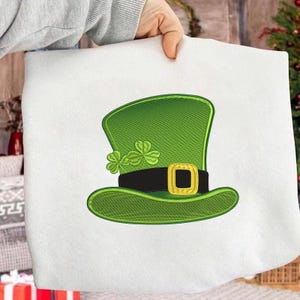 May include: A white fabric item featuring an embroidered green leprechaun hat with a black band and gold buckle. The hat is adorned with three-leaf clovers, a symbol of St. Patrick's Day.