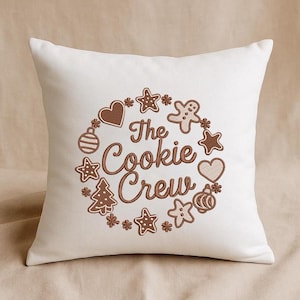 Op de afbeelding: Een vierkant, crèmekleurig kussen met een bruin geborduurd ontwerp. Het ontwerp toont de tekst "The Cookie Crew" omringd door peperkoekmannetjes, sterren, harten en kerstboomvormen. Het kussen is een decoratief item.