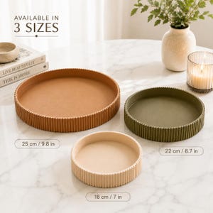 May include: Three round trays with ribbed sides in tan, olive green, and cream. The trays are labelled with diameters of 25 cm / 9.8 in, 22 cm / 8.7 in, and 18 cm / 7 in. Text in the image states AVAILABLE IN 3 SIZES, LIVE BEAUTIFULLY, and THIS IS HOME.