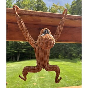 May include: A hanging monkey figurine crafted from woven light brown wicker material. The monkey features long arms hooked over a wooden beam, a rounded head with black and white plastic eyes, and a curved body with bent legs and feet.