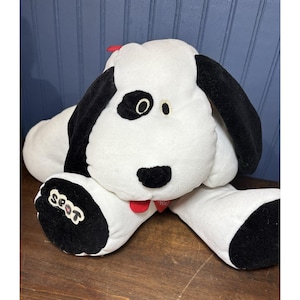 Peut inclure: Un jouet en peluche en forme de chien blanc avec des taches, des oreilles et des pattes noires. Le chien a une langue rouge et une étiquette en forme de cœur rouge. Le mot "seat" est sur une patte.