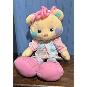 May include: A plush, yellow bear doll with pink accents. The bear has a pink yarn hairstyle, blue and purple heart-shaped accents, and a dress with the text "You're so sweet!" on the front. The doll has large pink feet.