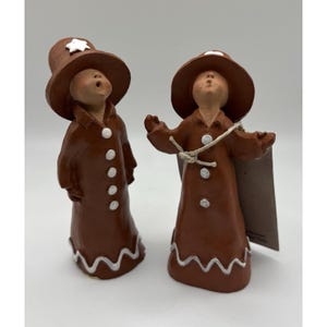 May include: Two brown gingerbread figurines with white accents. Each figure wears a hat and coat with white buttons and a wavy white trim. One hat has a white star. The figures are standing and looking upwards.
