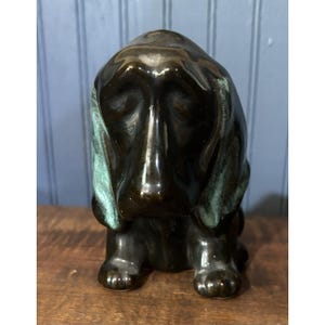 May include: A glossy dark brown ceramic figurine of a hound dog with long, drooping ears. The ears feature a distinct verdigris green patina finish, contrasting with the smooth, reflective dark brown glaze covering the rest of the dog's body.
